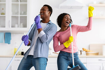 Happy african american lovers singing songs and dancing while house-keeping in white kitchen. Positive young black man and woman cleaning house together and having fun, copy space