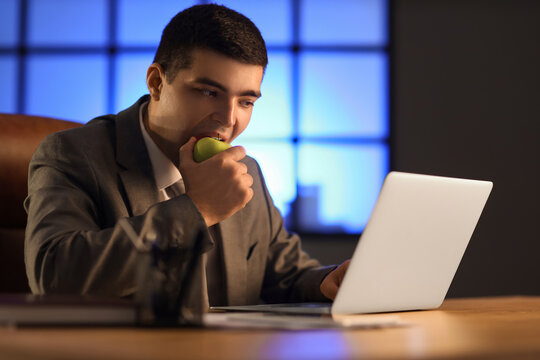 Young businessman eating apple in office at night