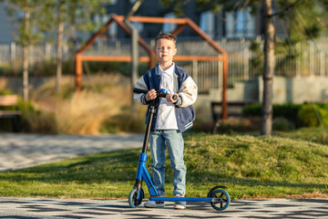 Child little boy stands with scooter on the sidewalk in a city park and looks at camera. Active...