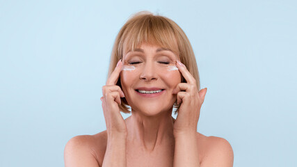 Mature beauty concept. Lovely senior woman applying face cream under her eyes over blue studio background, banner design with empty space. Older lady taking care of her skin