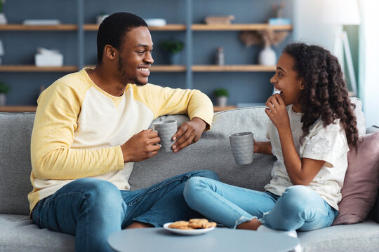 Joyful black father and daughter drinking tea with cookies in living room, sitting on couch and talking, copy space. Happy african american daddy and kid with mugs and biscuits having snack
