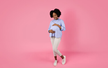 Positive young black pregnant lady in casual touches big belly, feeling movement, enjoy motherhood...