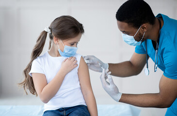 Young black doctor making covid-19 vaccine injection to little girl in face mask at hospital. Child...