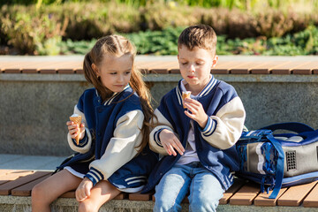 Two student children with backpacks, little boy and girl sitting an a wooden bench in the park, eating cold ice cream after school lessons. Concept of friendship and family relationship.