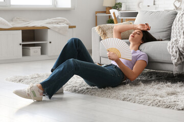 Young pretty woman with hand fan suffering from heat and lying on floor in living room