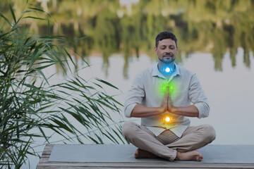 Man meditating in Lotus pose on pier near river. Glowing bubbles representing chakras