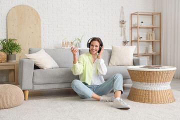 Smiling young woman in headphones with air conditioner remote control sitting near sofa in living...