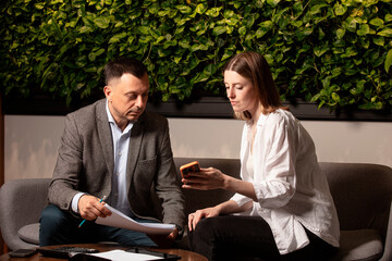 Two colleagues analyze data on a smartphone during a serious business meeting. Modern cozy office, greenery on the wall, documents and calculator on the table add realism. Preparation before the deal.