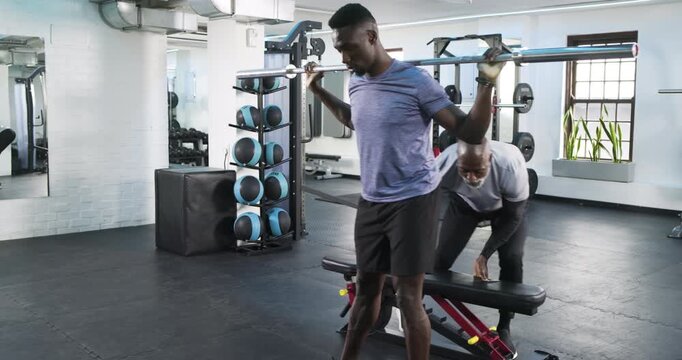 African American male trainer placing bench at gym while trainee balancing barbell for split squat