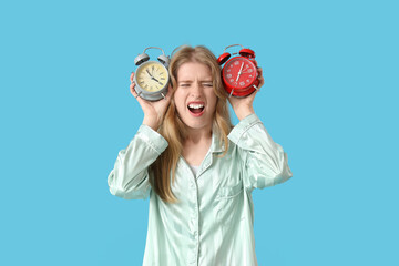 Stressed young woman in pajamas with alarm clocks suffering from insomnia on blue background