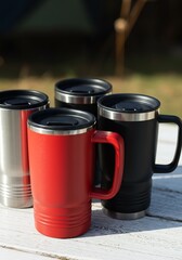 Red, black, and silver stainless steel travel mugs on rustic white wood.
