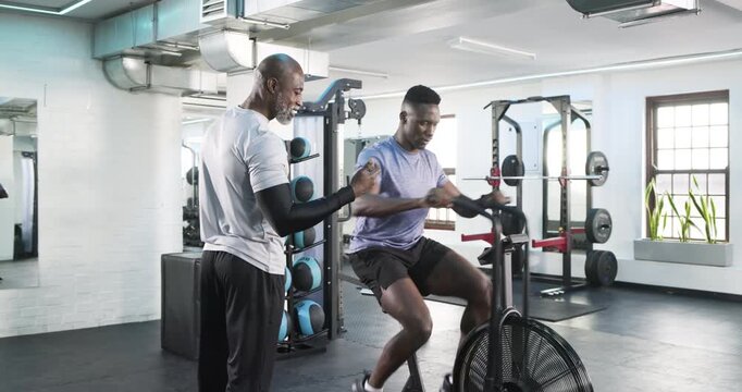 African American coach and trainee pedaling bike in gym on coach entry adjusting posture for form