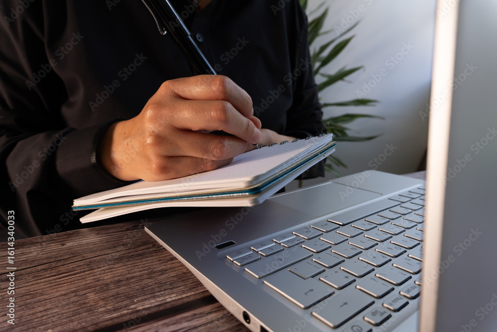 Poster business person sitting in front of a computer while writing something down in a notebook