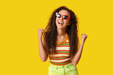 Happy young African-American woman in sunglasses showing yes gesture on yellow background