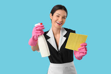 Beautiful young chambermaid with bottle of detergent and rag on blue background