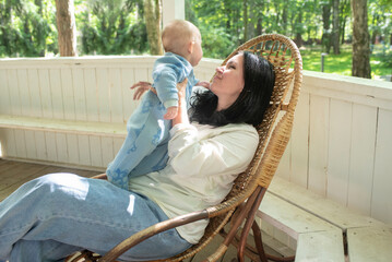 Mother with baby son sitting on rocking chair in white gazebo. Parenthood after 40, woman is 42 years old, baby is 6 months old.
