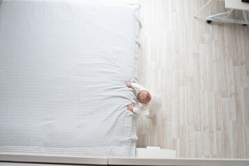 A small child, a baby in white clothes, holds on to a large parental bed and stands in a white room. View from above. The infant learned to stand by holding on to a support.