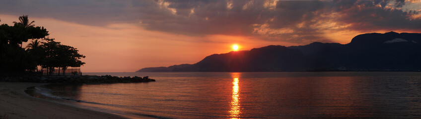 An intense orange sun setting behind a dark mountain range, creating a strong contrast of silhouettes on the horizon.