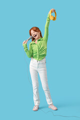 Teenage redhead girl with microphone and headphones singing karaoke on blue background