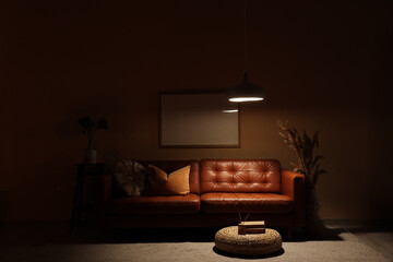 Interior of dark living room with brown sofa, blank frame and glowing lamp at night