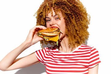 Young woman with curly hair enjoying a delicious burger, wearing a striped red and white shirt, vibrant colors highlight a joyful eating concept