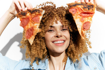 Happy young woman with curly hair holding two slices of pizza, showcasing a fun and playful attitude against a bright background