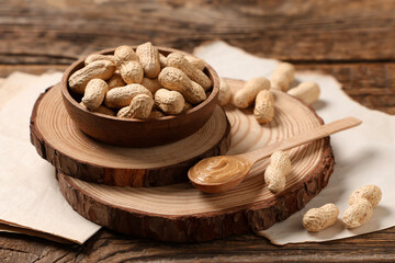 Bowl with unpeeled peanuts and spoon of nut butter on wooden background, closeup