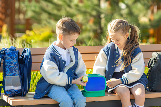 Two children, boy and girl eating lunch in school yard during break, sitting on the bench. Share the food, talking, laughing, resting outdoor. Back to school routine.