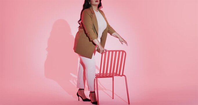 Pausing, woman in blazer walking to red chair in pink studio, straddling seat for relaxed pose