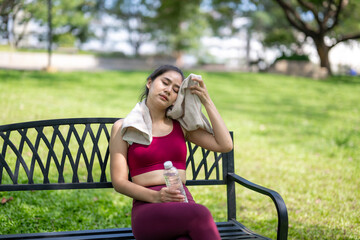 A woman sits on a park bench, wiping her forehead with a towel