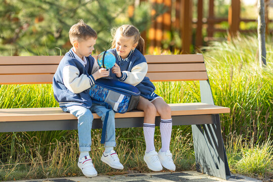 Two happy school children with backpacks, little boy and girl sitting on bench in park, exploring a globe together, learning geography, dreaming on travel. High quality photo. Back to school concept