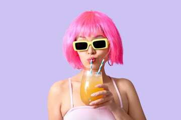 Pretty young woman in swimsuit and sunglasses drinking glass of orange juice with straws on lilac...