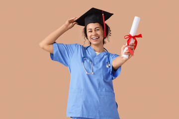 Female African-American medical student in graduation hat with diploma on beige background