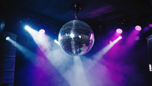 Sparkling disco ball hanging from the ceiling illuminated by vibrant purple, blue, and white stage lights in a smoky dark club atmosphere, evoking a festive nightlife and party dance floor mood.