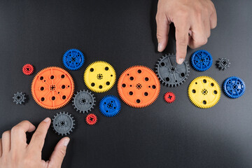 Colorful plastic gear wheels with human hands on black background, concept of teamwork and mechanics