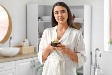 Pretty young woman holding bowl with sea salt, bath bomb and plumeria flower in bathroom