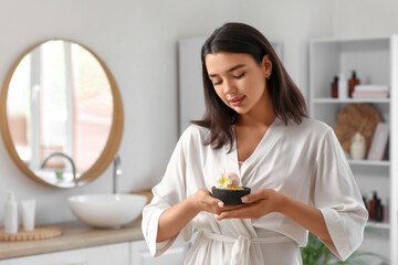 Pretty young woman holding bowl with sea salt, bath bomb and plumeria flower in bathroom