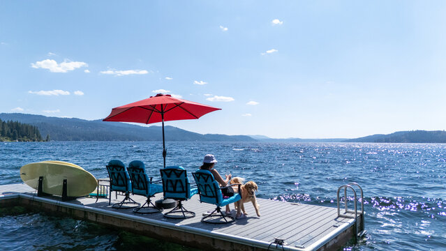 Woman sitting on a dock overlooking a Lake. She is sitting on a blue chair under a red umbrella on a sunny day while on vacation relaxing. 