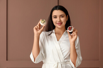 Pretty young woman in bathrobe with bath bombs on brown background
