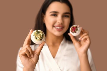 Pretty young woman in bathrobe with bath bombs on brown background