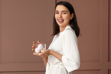Happy young woman in bathrobe holding bowl with bath bombs on brown background