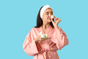 Pretty young woman in bathrobe sniffing bath bombs on blue background