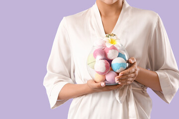 Pretty young woman in bathrobe holding bowl with bath bombs and plumeria flower on lilac background