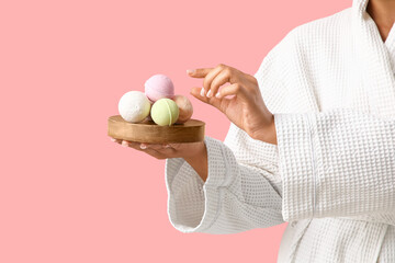 Pretty young woman in bathrobe with bath bombs on pink background