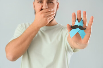 Young man with blue ribbon and paper mustache on light background, closeup. Prostate cancer...