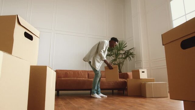 An African American man carries a box towards the bright living area of a new apartment. The space features a couch, while packing boxes are scattered around him.