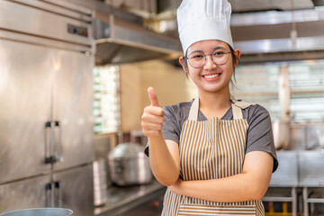 Young female chef smiling and showing a thumbs-up gesture in a professional kitchen environment. She wears a striped apron and a white chef hat, conveying positivity, approval, and confidence.