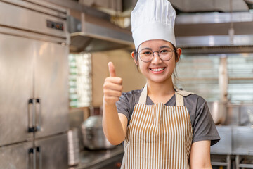 Young female chef smiling and showing a thumbs-up gesture in a professional kitchen environment. She wears a striped apron and a white chef hat, conveying positivity, approval, and confidence.