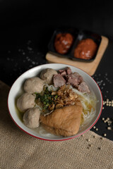 A delicious bowl of Indonesian Bakso soup with beef meatballs and fried tofu, served on a dark rustic background with chili sauce and burlap.