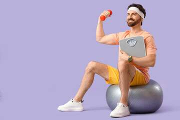 Sporty young man with weight scales and dumbbell training on fitball against lilac background